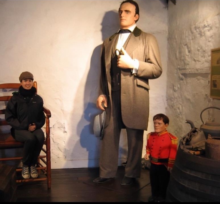 Museum photograph: the life-size Angus MacAskill statue in a gray period suit towers at center, holding his hat, while a smiling woman in a cap sits in a wooden chair beside him, her head reaching about his waist. A small figure of General Tom Thumb in a red uniform stands at the statue's knee beside a barrel and kettle display.