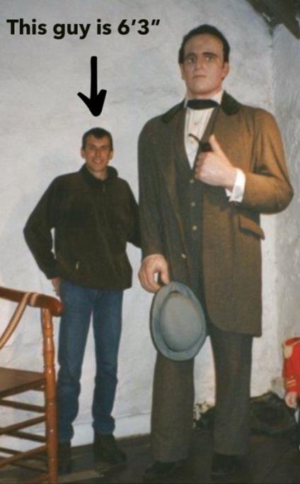 Museum photograph: a man stands beside the Angus MacAskill statue with an annotation reading 'This guy is 6\'3"' and an arrow pointing to him. The top of his head reaches roughly the statue's chest; the statue, in period suit with a bow tie, holds its hat at hip height.